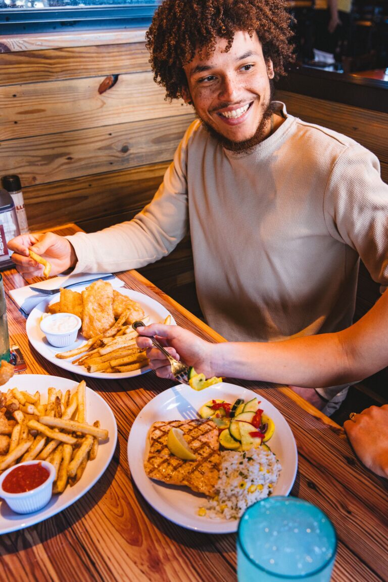 Smiling man at Miller's Ale House enjoying grilled fish, rice, vegetables, and fries on a wooden table.